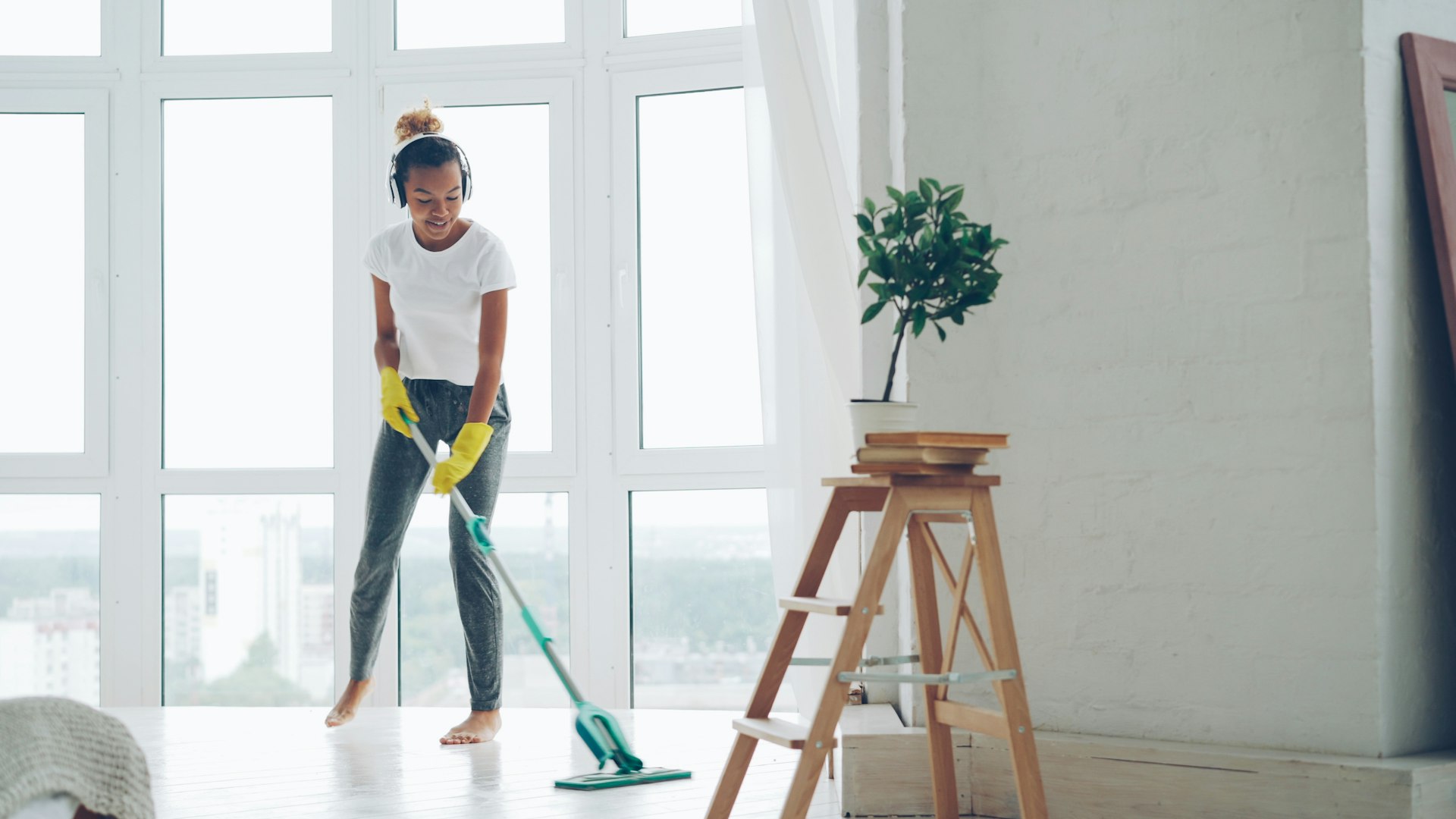A woman in yellow gloves is cleaning a large glass window with a cloth, smiling as she works. In the background, two people are engaged in cleaning tasks within a modern office space. One person is using a vacuum cleaner, while the other is at a desk. The setting has large windows and a dark backdrop.